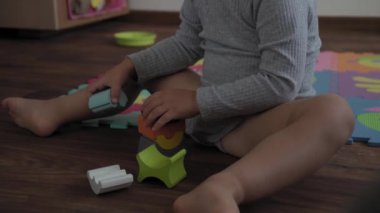 Çocukluk, aile, izolasyon konsepti. Küçük, şirin bir çocuk yerde renkli yumuşak bir yapboz üzerine ahşap piramit inşa ediyor. Çocuk evde motor becerileri için tek başına eğitim oyunları oynuyor..