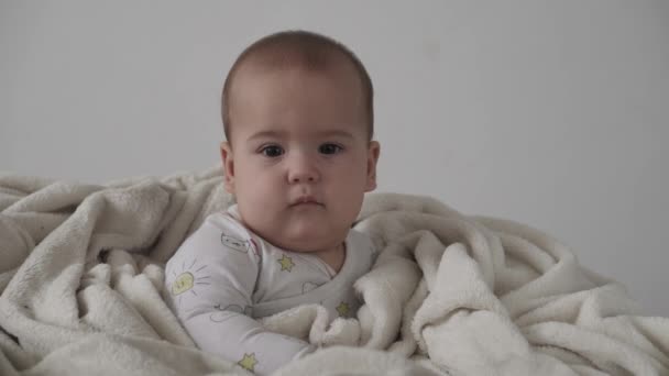 Βρέφος, έννοια της παιδικής ηλικίας - γκρο πλαν χαμογελαστό χαρούμενο αστείο καστανομάτικο παχουλό πρόσωπο του νεογέννητου παιδιού ξύπνιοι. χωρίς δόντια 7 μήνες μωρά γκριμάτσες, κλείνει τα μάτια κάθονται σε μαλακό κρεβάτι τυλιγμένο σε ζεστή κουβέρτα στο σπίτι — Αρχείο Βίντεο
