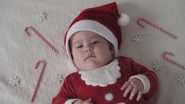 Joyeux noël, bonne année, nourrissons, enfance, concept de vacances - gros plan souriant 7 mois nouveau-né drôle dans santa claus chapeau, costume rouge jouer sur blanc doux lit jouer lécher bonbons de Noël doux — Video