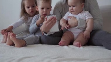 Çocukluk, annelik, kardeşlik, sohbet konsepti - Tasasız genç anne küçük çocuklarla çizgi film izler, yeni doğmuş bebek yatakta eğlenir, telefonda video konferans ile mutlu aile sohbetleri