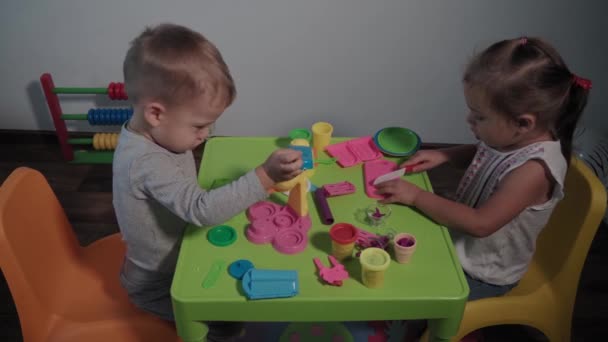 Medycyna, rodzina, gra, dzieciństwo, koncepcje sztuki - dwoje szczęśliwych małych dzieci bawi się kolorowymi lodami w fabryce plastycyny siedząc przy stole w domu podczas kwarantanny. Rodzeństwo dobrze się razem uczy. — Wideo stockowe