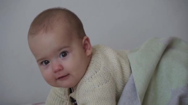 Säugling, Kindheit, Emotionskonzept - Nahaufnahme des lächelnden Gesichts eines braunäugigen, pummeligen, schläfrigen Babys von 7 Monaten in warmem Pullover, das gerade auf den Knien schaukelt, bedeckt mit einer warmen Decke auf einem weißen Bett zu Hause — Stockvideo