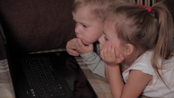 Famiglia, gioco, quarantena, concetti di infanzia - Due bambini felici che guardano cartoni animati sul computer portatile sdraiati sul divano marrone a casa durante la quarantena. Bambini fratelli fratello, sorella si divertono insieme — Video Stock