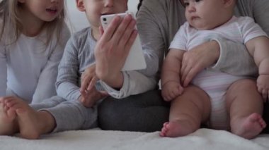 Çocukluk, annelik, kardeşlik, sohbet konsepti - Tasasız genç anne küçük çocuklarla çizgi film izler, yeni doğmuş bebek yatakta eğlenir, telefonda video konferans ile mutlu aile sohbetleri