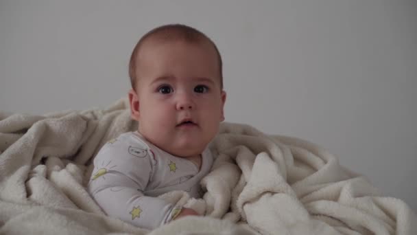 Младенческая, детская концепция - крупный план улыбающегося веселого каштанового пухлого лица новорожденного ребенка, бодрствующего. Беззубик 7 месяцев детские гримасы, закрывает глаза сидит на мягкой кровати завернутый в теплое одеяло дома — стоковое видео