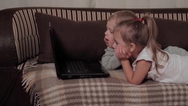 Famille, jeu, quarantaine, concepts d'enfance - Deux jeunes enfants heureux regardant des dessins animés sur ordinateur portable couché sur un canapé brun à la maison pendant la quarantaine. Enfants frères et sœurs frère, soeur s'amuser ensemble — Video