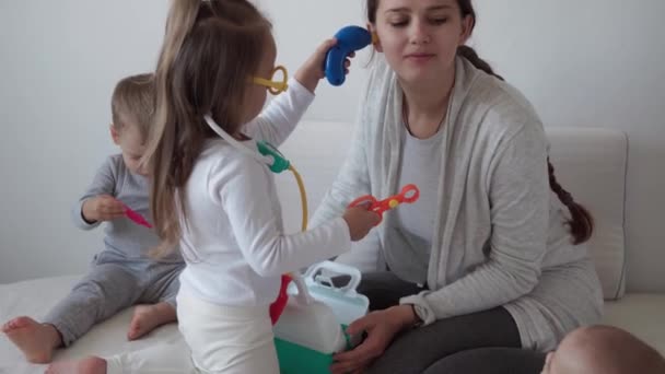 Medicina, familia, conceptos de juego: los niños pequeños y lúdicos concentrados usan anteojos médicos y usan estetoscopio. Fingir ser médico enfermera, dentista trata sonrisa mamá recién nacido bebé hermana dientes, sentarse en la cama — Vídeos de Stock