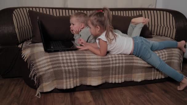 Familie, joc, carantină, concepte din copilărie - Doi copii mici fericiți care urmăresc desene animate pe laptop care stau pe canapea maro acasă în timpul carantinei. Copii frați frate, sora se distrează împreună — Videoclip de stoc