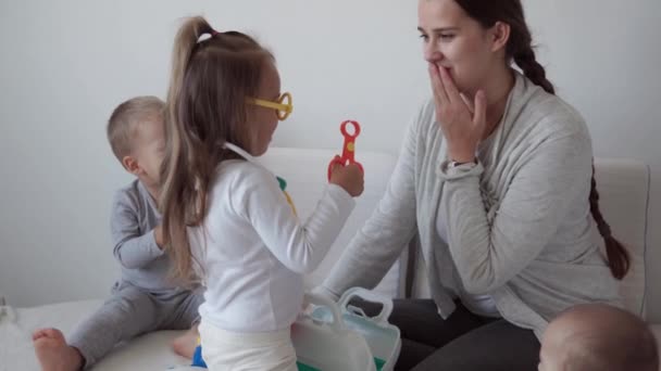 Medicin, familj, spel koncept - Koncentrera lekfulla små barn barn bära medicinska glasögon använder stetoskop. Låtsas vara läkare sjuksköterska, tandläkare behandlar leende mamma nyfödda lillasyster tänder, sitta på sängen — Stockvideo