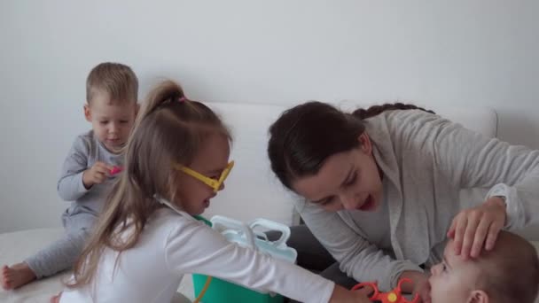 Medicin, familj, spel koncept - Koncentrera lekfulla små barn barn bära medicinska glasögon använder stetoskop. Låtsas vara läkare sjuksköterska, tandläkare behandlar leende mamma nyfödda lillasyster tänder, sitta på sängen — Stockvideo