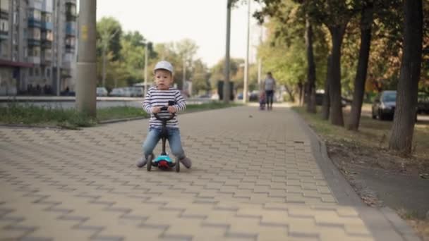 Childhood, family, vacation, transport concepts - preschool age slavic kid boy in cap ride push scooter in summer park near road trace, minor child express carefree, happiness enjoy leisure outdoors. — Stock Video