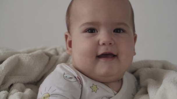Säugling, Kindheit Konzept - Nahaufnahme des lächelnden glücklich lustig braun-Augen pummelige Gesicht des neugeborenen Kindes wach. Zahnlos 7 Monate Baby Grimassen, schließt die Augen sitzen auf weichem Bett in warme Decke gewickelt zu Hause — Stockvideo