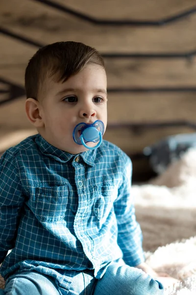 Enfance, famille, solitude concepts - Petit garçon noir d'âge préscolaire en vêtements bleus Perse apparence arabe de deux ans de nationalité mixte assis sur un lit léger doux avec sucette dans la bouche à la maison — Photo