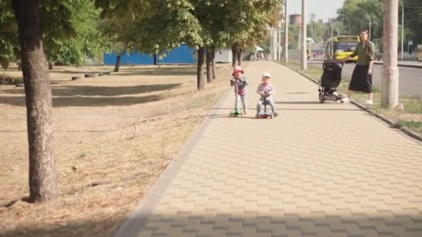 Kindheit, Familie, Urlaub, Verkehrskonzepte - slawische Kinder im Vorschulalter fahren Roller im Sommerpark nahe der Straße, fröhlich lächelnde minderjährige Kinder genießen die Freizeit im Freien. — Stockvideo