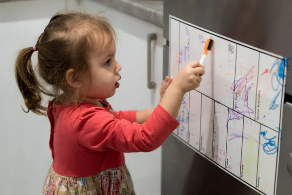 La infancia, el concepto de los lactantes - la niña pequeña rubia menor preescolar 2-3 años en el vestido dibuja con el marcador en la tabla de planear en el refrigerador en la cocina. Bebé niño ser creativo en la nevera con imanes — Foto de Stock