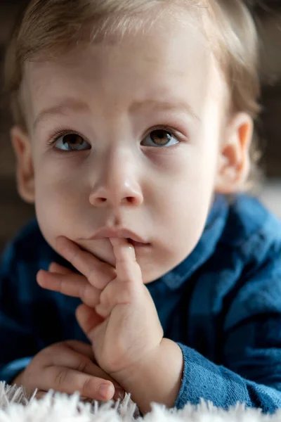 Childhood, infant, minor concepts - close up one black-eyed child baby boy is concentrated gnaws sucks fingers in mouth on bed. portrait of beautiful preschool Slavic kid in blue shirt at home indoors — Stock Photo, Image