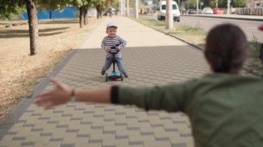 Çocukluk, aile, tatil, ulaşım kavramları. Sevimli anaokulu çocuğu yaz parkında scooter sürüyor. Annesinin kollarına yakın bir yerde.