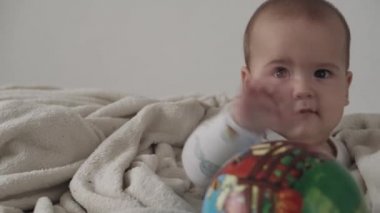 Bebek, çocukluk konsepti. Şirin, gülümseyen, komik kahverengi gözlü, tombul, yeni doğmuş çocuğun yakın çekimi. Dişsiz 7 aylık bebek evde sıcak battaniyeye sarılı beyaz yumuşak bir yatakta oturmuş topla oynuyor.