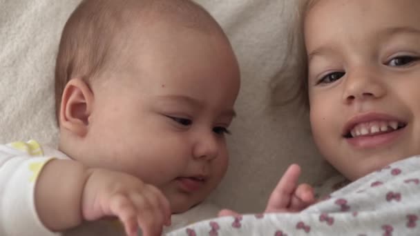 Infancia, infancia, familia, sueño, concepto de descanso: primer plano de dos alegres niños felices, un bebé recién nacido y una niña de 3-4 años se acuestan juntos en una cama beige y blanca. Las hermanas están jugando en el sofá en casa. — Vídeos de Stock