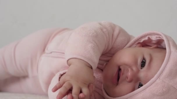 Spädbarn, barndomskoncept - närbild av leende glad fyllig rolig brunögd knubbig ansikte nyfödda barn vaken. tandlös 7 månaders baby grimaces, sluter ögonen ligger på mjuk säng i rosa gnagare fingrar på sängen — Stockvideo