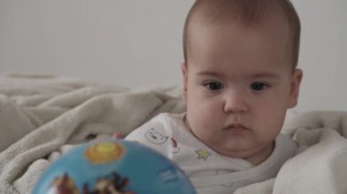Bebek, çocukluk konsepti. Şirin, gülümseyen, komik kahverengi gözlü, tombul, yeni doğmuş çocuğun yakın çekimi. Dişsiz 7 aylık bebek evde sıcak battaniyeye sarılı beyaz yumuşak bir yatakta oturmuş topla oynuyor.