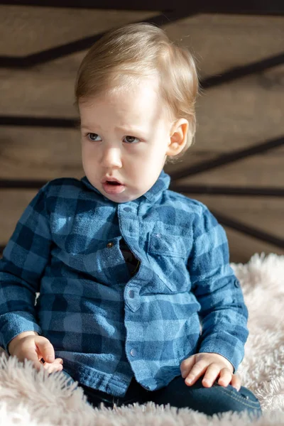 Infancia, bebé, conceptos menores - cerca de una rubia de ojos negros triste, niño ofendido bebé niño en la cama polvorienta suave en habitación soleada. retrato avergonzado preescolar niño eslavo en camisa en casa en interiores — Foto de Stock