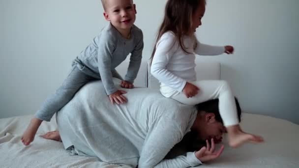 Childhood, motherhood, siblings concept - Carefree playful young mum, two little kids daughter, son have fun play on bed, happy cheerful family small child girl laugh tickles, kisses, hugs lying relax — Stock Video