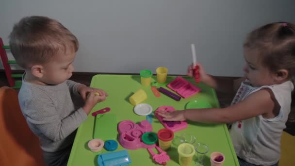 Medicina, familia, juego, infancia, conceptos de arte Dos niños pequeños y felices juegan con la fábrica de helados de plastilina de colores sentados en la mesa durante la cuarentena. Los hermanos niños se divierten estudiando juntos — Vídeos de Stock