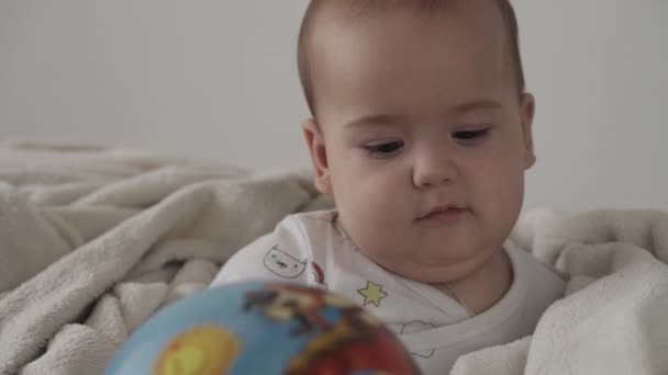 Infantil, conceito de infância - close-up de bonito sorrindo feliz engraçado de olhos castanhos gordinho recém-nascido criança acordada. toothless 7 meses bebê jogar com bola sentado na cama macia branca envolto em cobertor quente em casa — Vídeo de Stock