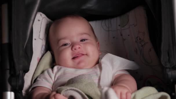 Zuigeling, kindertijd, emotie concept - close-up van glimlachend gezicht van bruin-ogige mollig pasgeboren wakker tandeloze baby 7 maanden oud kijken naar camera liggend in wit bodysuit met gekleurde strepen in kinderwagen — Stockvideo