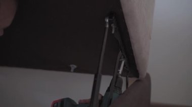 Yenileme, inşaat konsepti. Yakın plan, el kamerası çekimleri. Elinde matkap ve mobilyaları büken İngiliz anahtarı olan bir erkek eli. Adamın biri evdeki yatak odasında kanepe topluyor..