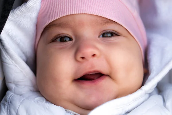 Младенца, детство, эмоциональная концепция - крупный план милого улыбающегося портрета с карими глазами пухлого новорожденного, бодрствующего беззубого ребенка 6-9 месяцев назад, смотрящего на камеру, лежащую в белой куртке, розовой шляпе в коляске — стоковое фото