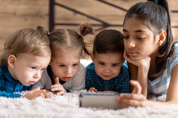 Vänskap, barndom, teknik koncept - 4 små barn baby av olika nationaliteter persiska och slaviska utseende titta på tecknad film på smartphone på sängen. barn förskola tala genom videokonferens — Stockfoto