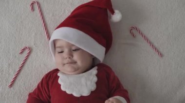 Mutlu Noeller, mutlu yıllar, bebekler, çocukluk, tatil konsepti - yakın plan gülümsemesi Noel Baba şapkalı komik bebek, kırmızı beden beyaz yumuşak yatakta oynuyor tatlı Noel şekerini yala