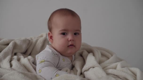 Criança, conceito de infância - close-up de sorrir feliz rosto gordinho engraçado de olhos castanhos de criança recém-nascida acordada. toothless 7 meses bebê grimaces, fecha os olhos sentar-se em cama macia envolto em cobertor quente em casa — Vídeo de Stock