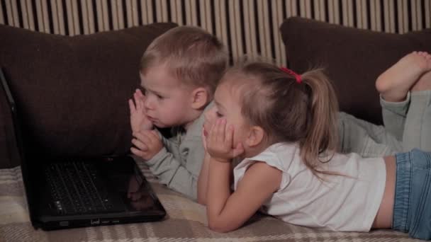 Familj, spel, karantän, barndom koncept - Två glada små barn tittar på karikatyrer på laptop ligger på brun soffa hemma under karantän. Barn syskon bror, syster ha kul tillsammans — Stockvideo