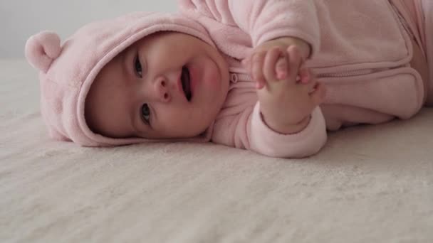 Criança, conceito de infância - close-up de sorrir feliz gordo rosto gordinho de olhos castanhos engraçado de criança recém-nascida acordada. toothless 7 meses bebê grimaces, fecha os olhos mentira em cama macia em rosa rói dedos na cama — Vídeo de Stock