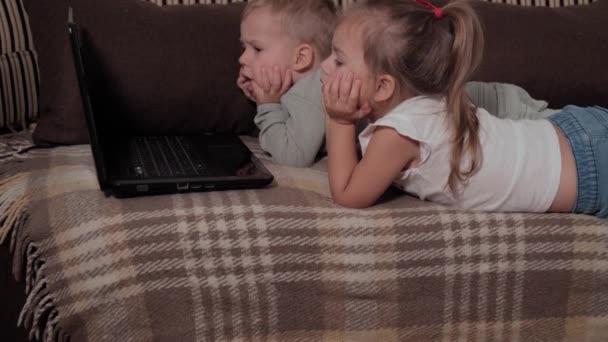 Famille, jeu, quarantaine, concepts d'enfance - Deux jeunes enfants heureux regardant des dessins animés sur ordinateur portable couché sur un canapé brun à la maison pendant la quarantaine. Enfants frères et sœurs frère, soeur s'amuser ensemble — Video