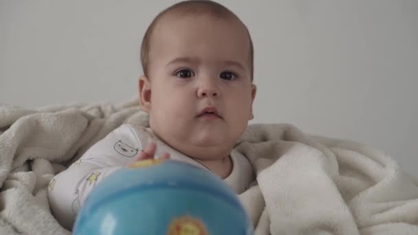 Νήπιο, παιδική έννοια - γκρο πλαν χαριτωμένο χαμογελαστό χαρούμενο αστείο καστανομάτικο παχουλό νεογέννητο παιδί ξύπνιο. χωρίς δόντια 7 μήνες παιχνίδι μωρό με μπάλα κάθεται σε λευκό μαλακό κρεβάτι τυλιγμένο σε ζεστή κουβέρτα στο σπίτι — Αρχείο Βίντεο