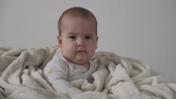 Spädbarn, barndomskoncept - närbild av leende glad rolig brunögd knubbig ansikte nyfödda barn vaken. tandlös 7 månader baby grimaces, sluter ögonen sitta på mjuk säng insvept i varm filt hemma — Stockvideo