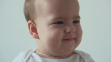 Bebek, çocukluk konsepti. Gülümseyen mutlu kahverengi gözlü, tombul çocuk uyandı. Neşesiz, dişsiz, 7 aylık bebek buruşuklukları, gözlerini kapatır evdeki beyaz arka planda yumuşak yatağa oturur.