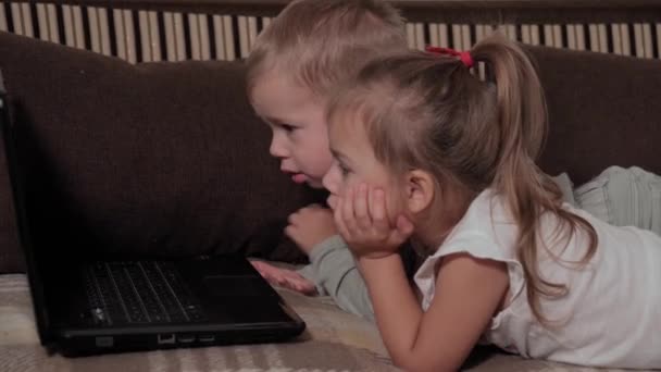 Familie, joc, carantină, concepte din copilărie - închideți doi copii fericiți care urmăresc desene animate pe laptopul care stă pe canapea maro acasă în timpul carantinei. Copii frați frate, sora se distrează — Videoclip de stoc