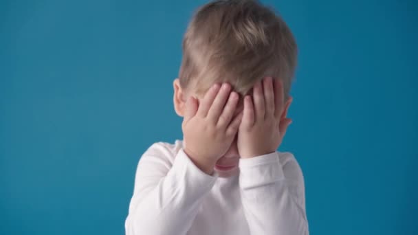 Estúdio tiro, felicidade, conceitos de infância - feliz, sorrindo, criança alegre pré-escolar menor Menino de criança posando olhar para a câmera, mostra caretas, obter olhos de cobertura com raiva, orelhas bater palmas no fundo azul. — Vídeo de Stock