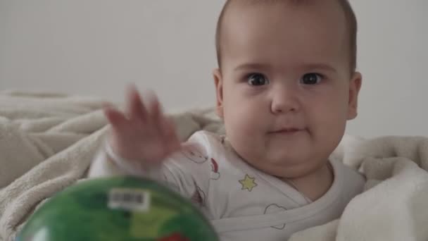 Νήπιο, παιδική έννοια - γκρο πλαν χαριτωμένο χαμογελαστό χαρούμενο αστείο καστανομάτικο παχουλό νεογέννητο παιδί ξύπνιο. χωρίς δόντια 7 μήνες παιχνίδι μωρό με μπάλα κάθεται σε λευκό μαλακό κρεβάτι τυλιγμένο σε ζεστή κουβέρτα στο σπίτι — Αρχείο Βίντεο