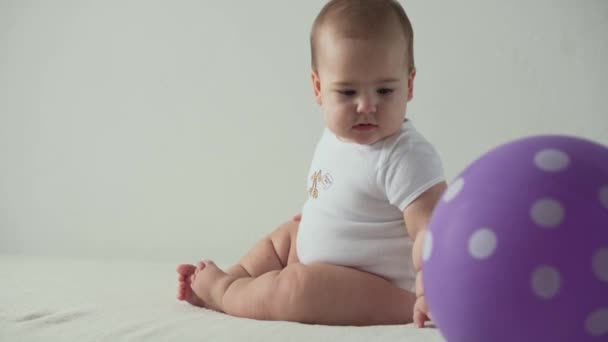 Ребенок, концепция детства - счастливый забавный playfull малыша 8 месяцев девочка играть с фиолетовым шариком на день рождения. Улыбающийся толстый активный бодрствующий ползает на коленях внутри дома на белом фоне — стоковое видео