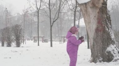 Kış, tatil, oyunlar, aile kavramları - mutlu anaokulu çocuğu kız çocuk evden kaçıp kartopu oynayarak eğleniyor. Kar fırtınası mevsiminde dışarıda, parkta, soğuk havada dönüyor..
