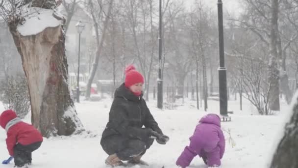 Inverno, férias, jogos, conceitos de família - Dois felizes crianças pré-escolares irmãos crianças vestidos com chapéus e luvas com a mãe jogando fazer bola de neve na queda de neve em tempo de estação fria no parque ao ar livre — Vídeo de Stock