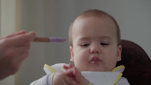 Cibo, nutrizione, concetti infantili - La mamma nutre il bambino con un cucchiaio di gomma. Carino non bambino affamato vestito in bavaglino si siede in sedia per bambini non vogliono mangiare piatto in cucina al chiuso. antipatia prima alimentazione — Video Stock