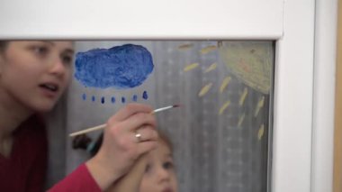 Çocukluk, annelik, yaratıcılık, doğa, sanat, bahar, yaz konsepti - 3-4 yaşlarında küçük kızı olan genç bir annenin pencere camına çizimi. Kadın ve anaokulu kızı güneşi boyuyorlar.