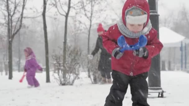 Зима, відпустка, ігри, сімейні концепції - Двоє щасливих дітей дошкільного віку брати і сестри одягнені в капелюхи і рукавиці з мамою, які грають, роблять снігову кулю в снігопаді в холодну погоду в парку на відкритому повітрі — стокове відео